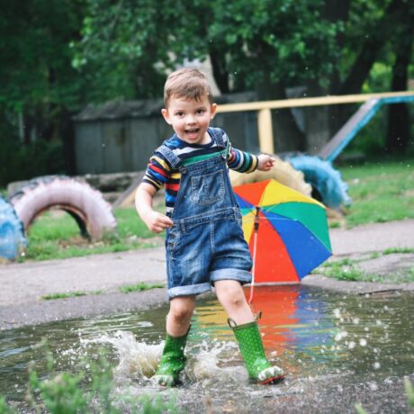 Băiețel, îmbrăcat cu o bluză cu dungi colorate și o salopetă din denim albastru, cu pantaloni scurți și cizme verzi, din cauciuc, care aleargă într-o baltă, în parc, ilustrând consecințele naturale în educația copiilor
