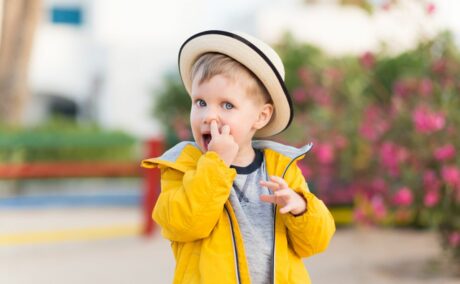 Băiețel în parc,care își bagă degetul arătător în nas, îmbrăcat cu o bluză gri, jachetă galbenă și are pe cap o pălărie bej, bordată cu bleumarin, ilustrând copilul își bagă degetul în nas