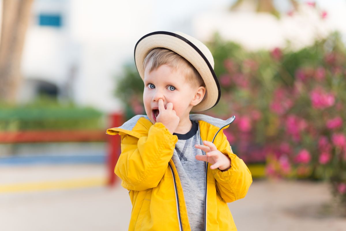 Băiețel în parc,care își bagă degetul arătător în nas, îmbrăcat cu o bluză gri, jachetă galbenă și are pe cap o pălărie bej, bordată cu bleumarin, ilustrând copilul își bagă degetul în nas