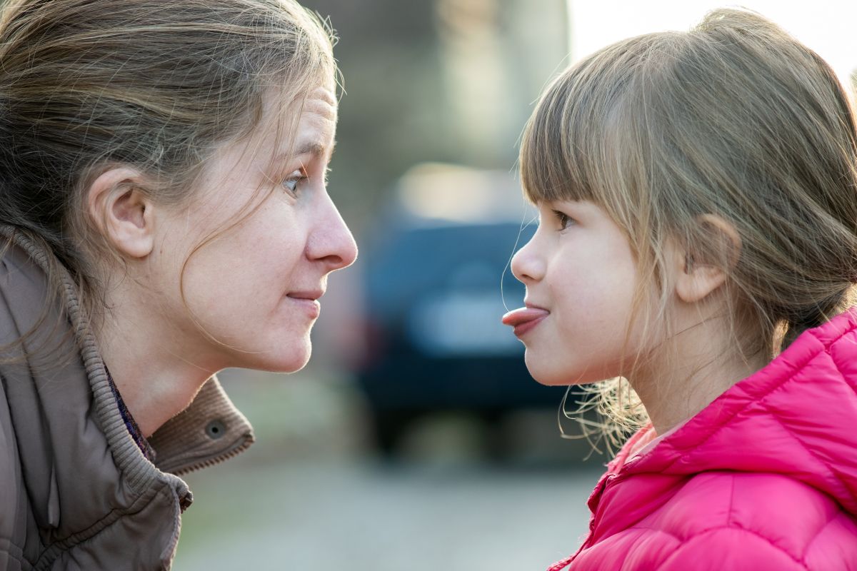 fetiță, îmbrăcată cu jachetă roz, care scoate limba la mama ei, în timp ce sunt pe stradă, ilustrând copilului nu-i pasă de consecințe