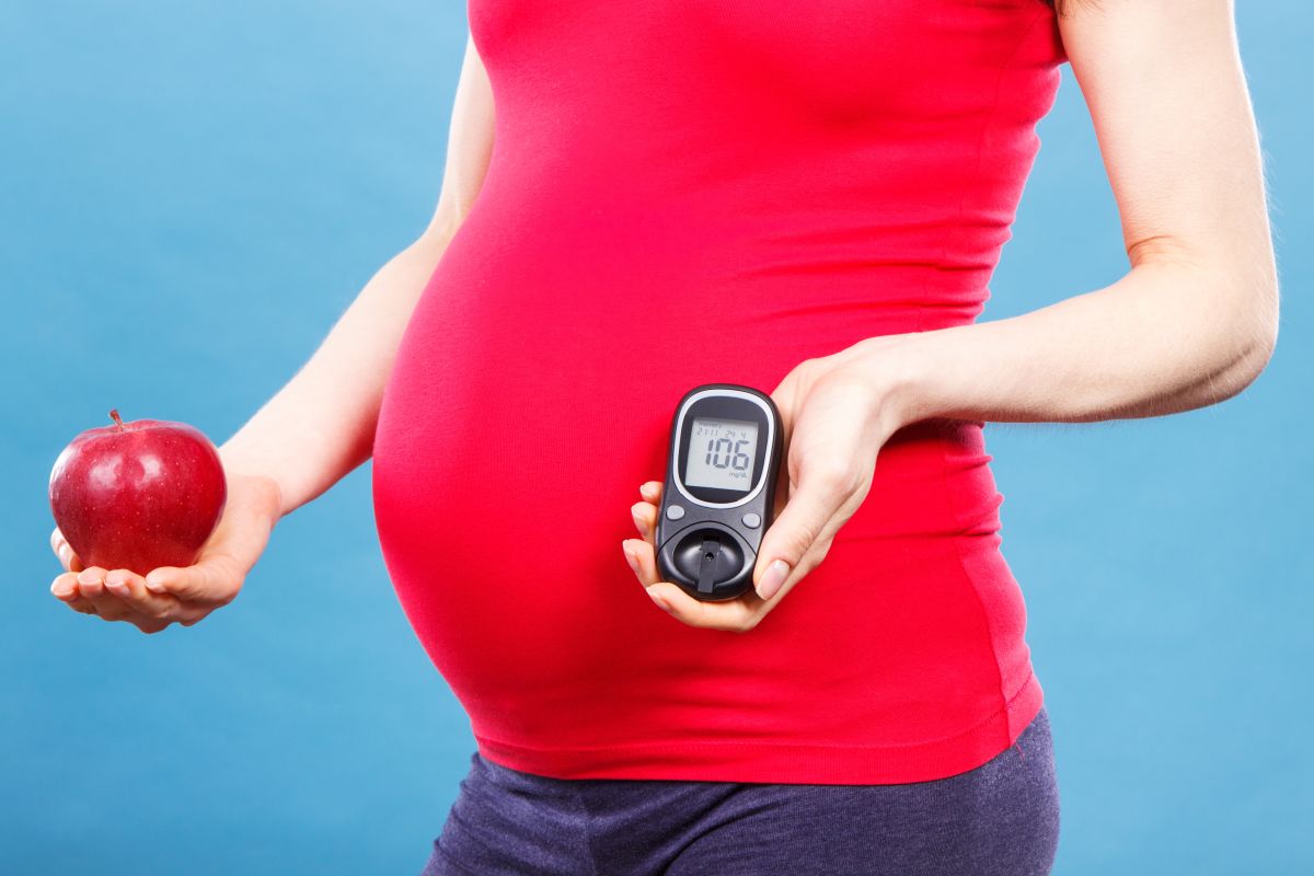 Femeie însărcinată, în tricou roșu, pe un fundal albastru, care ține într-o mână un măr roșu și în cealaltă un aparat de măsurare a glicemiei, ilustrând Cum afectează diabetul sănătatea reproductivă a femeii