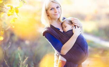Mămică blondă, îmrăcată cu o bluză albastră, care a ieșit în parc cu bebelușul pe care îl ține într-un wrap, ilustrând cum să alegi un sistem de purtare