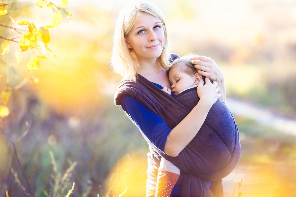 Mămică blondă, îmrăcată cu o bluză albastră, care a ieșit în parc cu bebelușul pe care îl ține într-un wrap, ilustrând cum să alegi un sistem de purtare