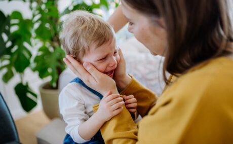 Mamă, îmbrăcată în galben, care ține fața băiețelului ei, care plânge, în palme, iar pe fundal, în cameră este o plantă cu frunze verzi, în ghiveci, ilustrând Cuvintele pe care nu ar trebui să le spui copilului tău