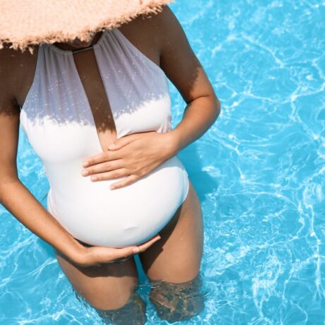 Femeie însărcinată, în piscină, îmbrăcată cu costum de baie alb, întreg, și pălărie din paie pe cap, ilustrând expunerea la soare în perioada sarcinii