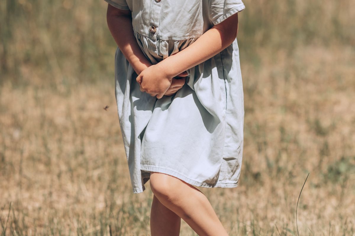 Fetiță, îmbrăcată cu o rochiță gri, care este pe un câmp și își ține picioarele apropiate și mâinile sub abdomen, ilustrând infecția urinară la copii