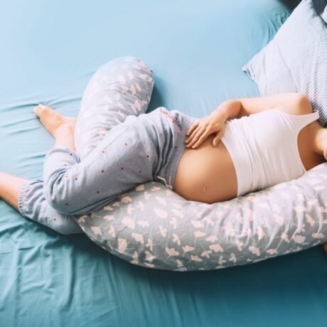 Femeie însărcinată, îmbrăcată cu un maiou alb și pantaloni de pijama gri cu inimioare roșii, care doarme pe un cearșaf bleu, cu o pernă de gravide între picioare, ilustrând insomnia în sarcină