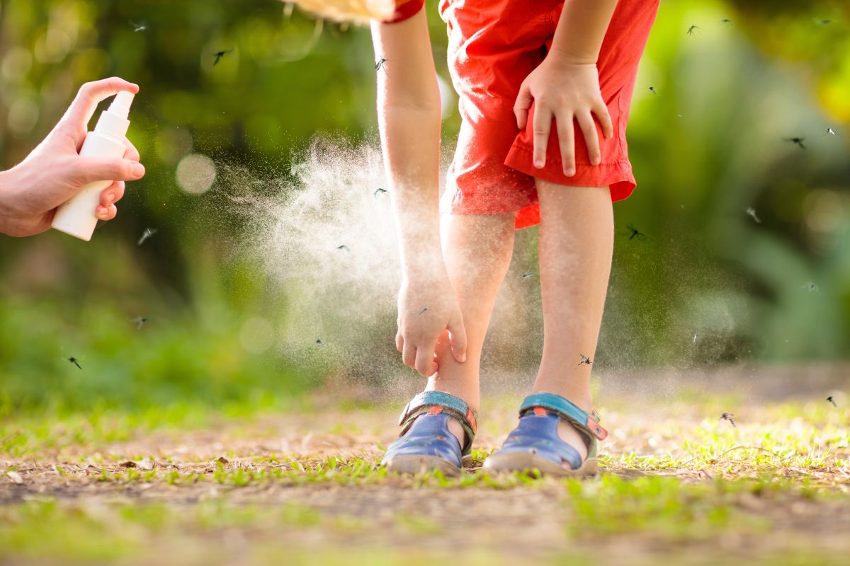 Băiețel, îmbrăcat cu pantaloni oranj și sandale albastre, care a ieși în parc și în jurul lui roiesc țânțarii, se carpină cu o mână și în imagine apare și mâna unui adult care ține un mână un spray, ilustrând înțepăturile de țânțari la copii