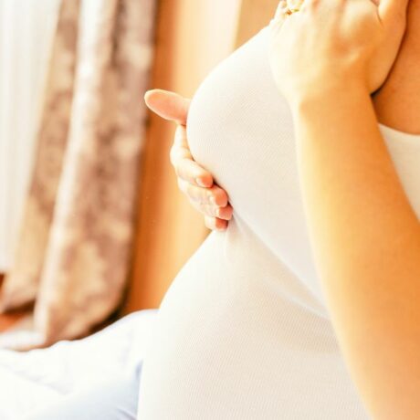Femeie însărcinată, îmbrăcată cu maiou alb, în dormitor care își palpează un sân cu mâinile, ilustrând modificările sânilor în timpul sarcinii