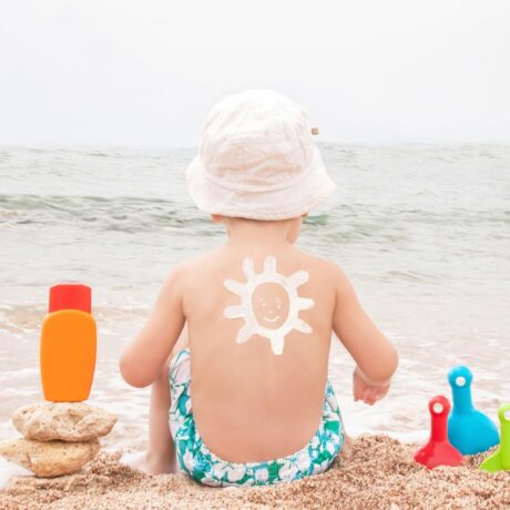 Băiețel care stă pe nisip, cu fața către mare, iar pe spate are desenat cu cremă albă un soare, având un slip cu flori albe, verzi și albastre, care are în stânga o cremă de protecție solară portocalie, așezată pe niște pietre, iar în partea cealaltă o lopețică roșie, una verde și una bleu, înfipte în nisip, ilustrând prevenirea arsurilor solare la copii