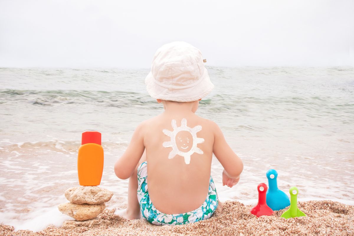 Băiețel care stă pe nisip, cu fața către mare, iar pe spate are desenat cu cremă albă un soare, având un slip cu flori albe, verzi și albastre, care are în stânga o cremă de protecție solară portocalie, așezată pe niște pietre, iar în partea cealaltă o lopețică roșie, una verde și una bleu, înfipte în nisip, ilustrând prevenirea arsurilor solare la copii