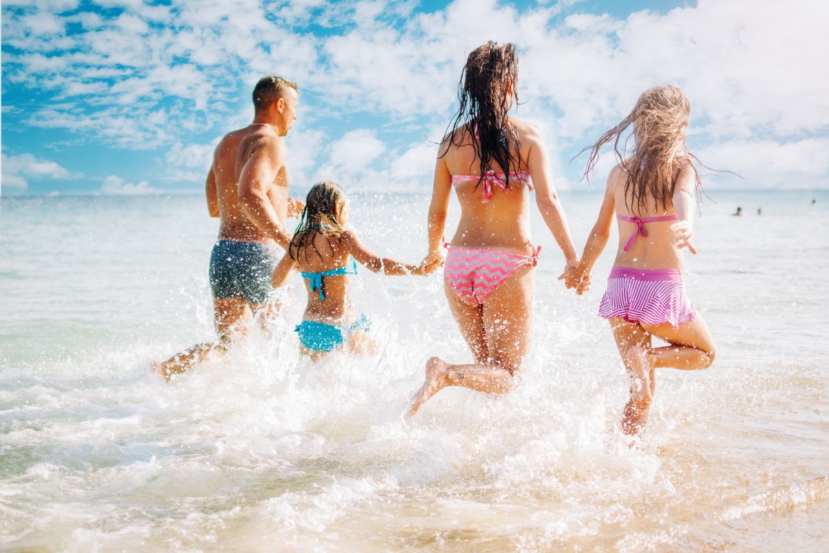 Familie în costume de baie colorate, fotografiată din spate, în timp ce aleargă prin mare, ilustrând radiațiile ultraviolete