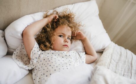 Fetiță, îmbrăcată cu o bluză de pijama albă cu buline mici bleumarin, care stă în pat, pe o pernă albă și este acoperită cu pilota și își ține mâinile în păr, ilustrând răsucirea părului la copii