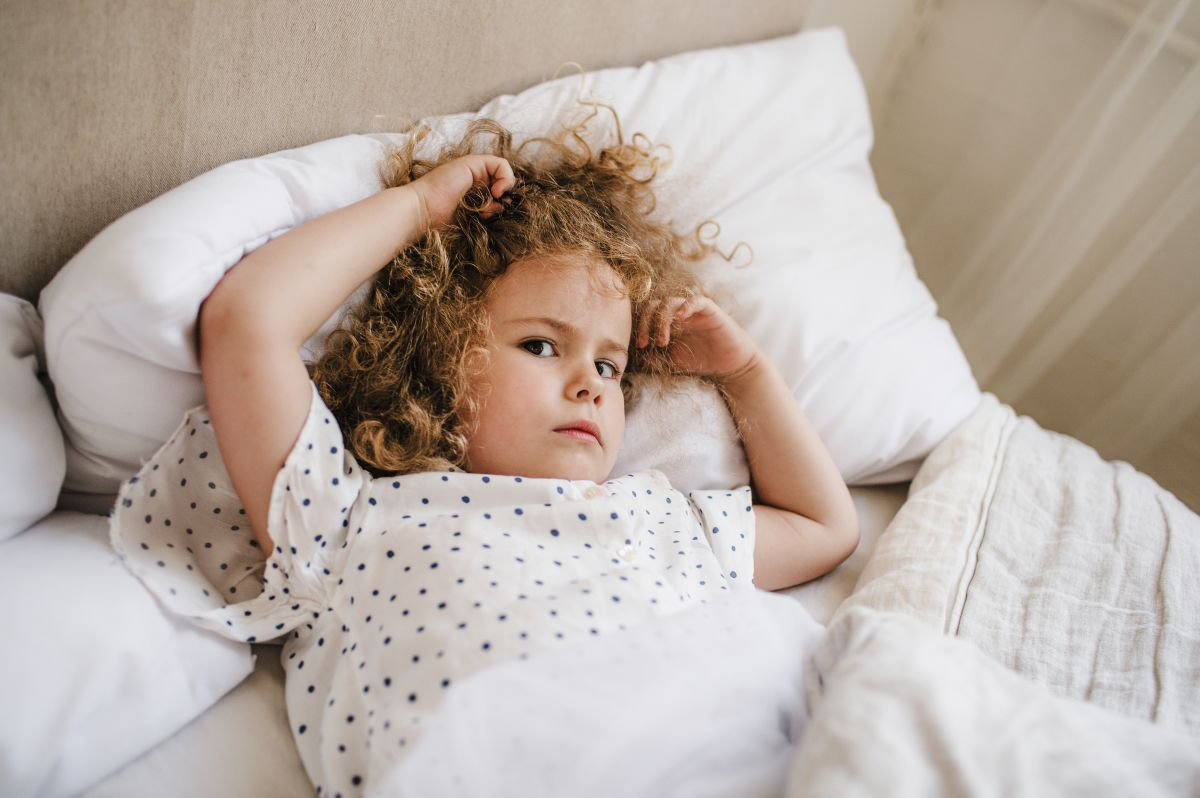 Fetiță, îmbrăcată cu o bluză de pijama albă cu buline mici bleumarin, care stă în pat, pe o pernă albă și este acoperită cu pilota și își ține mâinile în păr, ilustrând răsucirea părului la copii