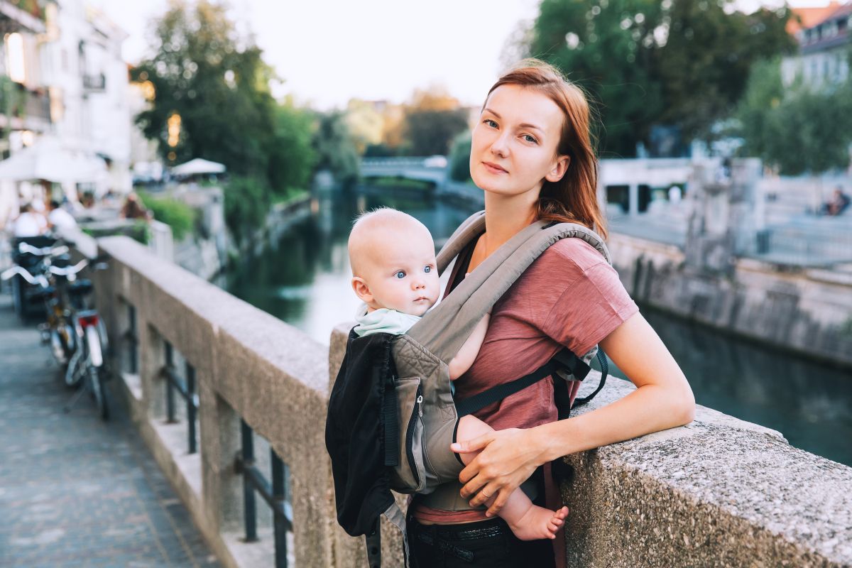 Mămică cu părul șaten, cu bebelușul în marsupiu, pe un pod