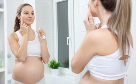 Femeie însărcinată, îmbrăcată cu o bustieră și colanți albi, în baie, care ține într-o mână un tub alb cu cremă și cu cealaltă o aplică, ilustrând tenul strălucitor în sarcină