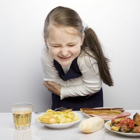 Fetiță, îmbrăcată cu bluză albă și rochie bleumarin pe deasupra, care se ține de stomac, iar în fața ei este o masă albă pe care sunt așezate o farfurie cu friptură, o farfurie cu mezeluri , una cu chips-uri , o chiflă și un pahar cu ceai, ilustrând toxiinfecția alimentară la copii