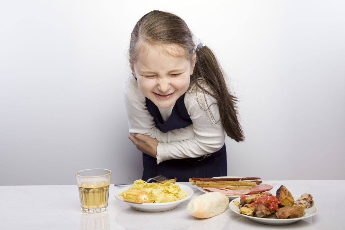 Fetiță, îmbrăcată cu bluză albă și rochie bleumarin pe deasupra, care se ține de stomac, iar în fața ei este o masă albă pe care sunt așezate o farfurie cu friptură, o farfurie cu mezeluri , una cu chips-uri , o chiflă și un pahar cu ceai, ilustrând toxiinfecția alimentară la copii