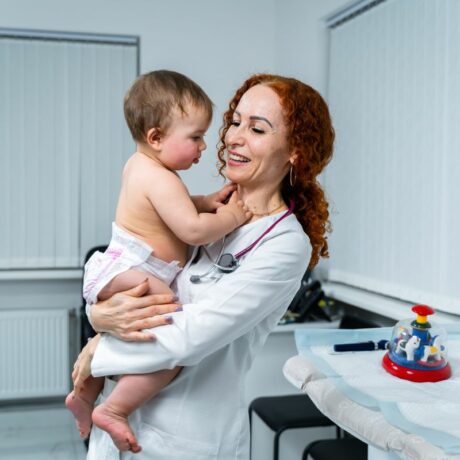Medic pediatru, în cabinet, care ține în brațe un bebeluș cu scutec, ilustrând vizita medicală de la 1 an a bebelușului