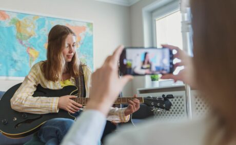 Adolescentă care cântă la chitară în camera ei, având în spate o hartă, iar mama ei o filmează cu telefonul, ilustrând 5 greșeli pe care le faci ca părinte în social media