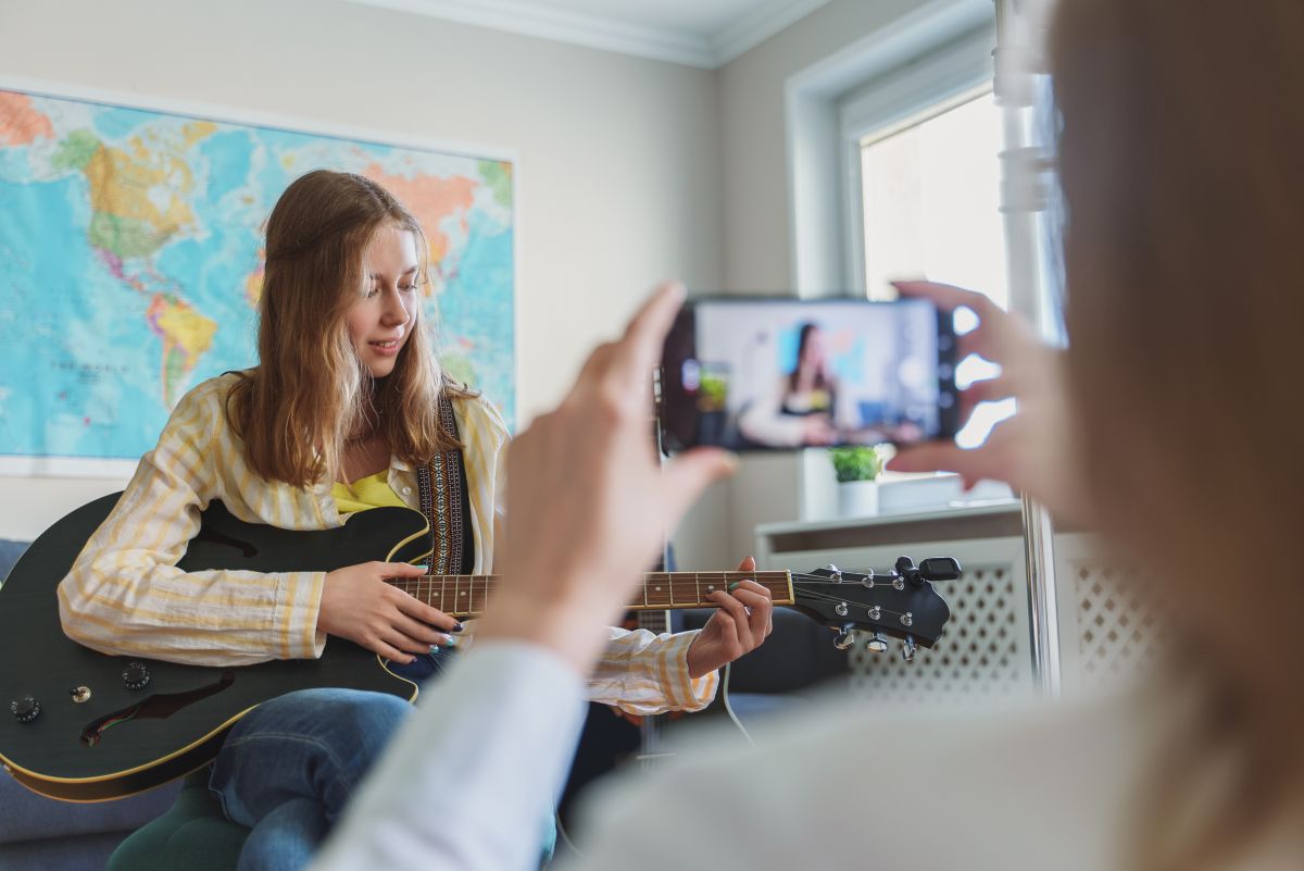 Adolescentă care cântă la chitară în camera ei, având în spate o hartă, iar mama ei o filmează cu telefonul, ilustrând 5 greșeli pe care le faci ca părinte în social media