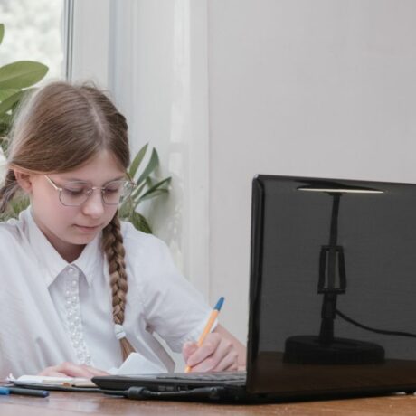 Fetiță, îmbrăcată cu cămașă albă, care stă la birou și scrie pe caiet cu un pix galben, având un laptop negru deschis în față, iar în spate o plantă verde, ilustrând autodisciplina la copii