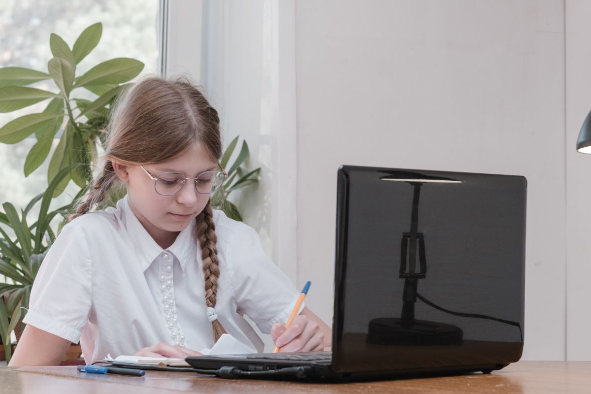 Fetiță, îmbrăcată cu cămașă albă, care stă la birou și scrie pe caiet cu un pix galben, având un laptop negru deschis în față, iar în spate o plantă verde, ilustrând autodisciplina la copii