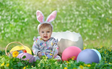 Bebeluș care stă pe o pajiște cu iarbă și flori galbene, având pe cap nisște urechi pufoase de iepuraș, alb cu roz, și lângă el un coș și niște ouă mari roz și albastre și o jumătate de coajă de ou albă, iar în maini ține câte un ou albastru, ilustrând bebelușii născuți în aprilie