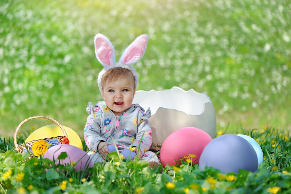 Bebeluș care stă pe o pajiște cu iarbă și flori galbene, având pe cap nisște urechi pufoase de iepuraș, alb cu roz, și lângă el un coș și niște ouă mari roz și albastre și o jumătate de coajă de ou albă, iar în maini ține câte un ou albastru, ilustrând bebelușii născuți în aprilie