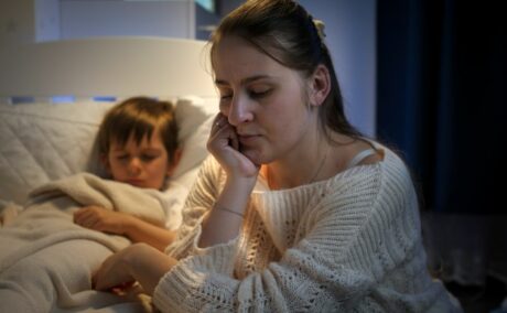 Mamă, îmbrăcată cu un pulover alb, care stă lângă patul copilului ei, care încearcă să doarmă, fiind acopit cu o pilotă, ilustrând copiii nu vor să meargă culcare