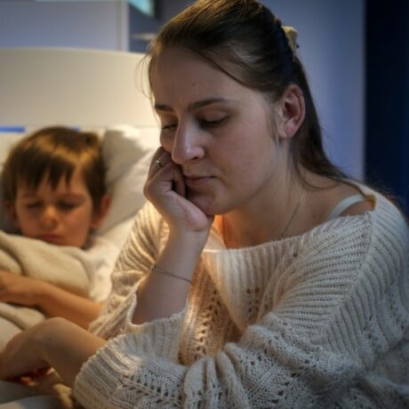 Mamă, îmbrăcată cu un pulover alb, care stă lângă patul copilului ei, care încearcă să doarmă, fiind acopit cu o pilotă, ilustrând copiii nu vor să meargă culcare