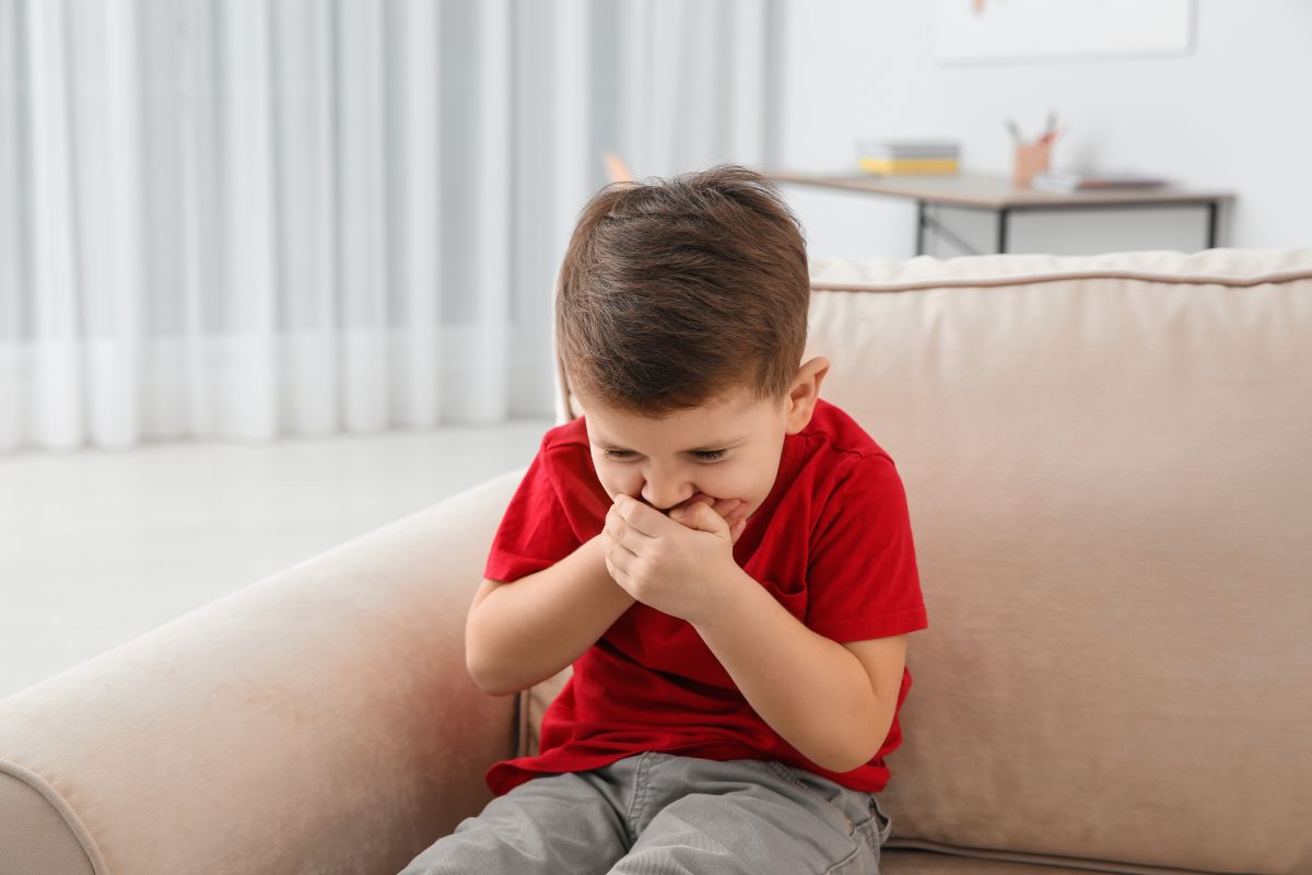 Copil, îmbrăcat cu un tricou roșu și pantaloni gri, care stă așezat pe o canapea bej și se ține cu ambele mâini de gură, ilustrând copilul vomită după ce i-ai dat medicamentul