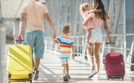 Familie, compusă din tată, care ține de mână un băiețel și în cealaltă mână ține un troller galben și mama, care ține în mână un troller rubiniu și o fetiță în brațe, toți se îndreaptă spre aeroport, ilustrând cum să eviți germenii când călătorești cu copiii