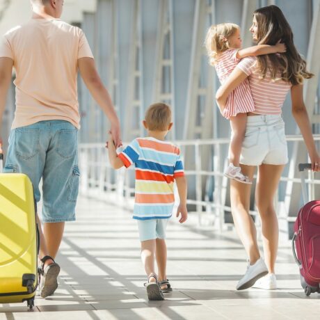 Familie, compusă din tată, care ține de mână un băiețel și în cealaltă mână ține un troller galben și mama, care ține în mână un troller rubiniu și o fetiță în brațe, toți se îndreaptă spre aeroport, ilustrând cum să eviți germenii când călătorești cu copiii