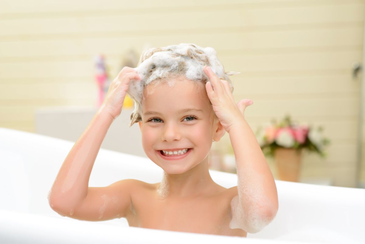 Fetiță, care stă într-o cadă albă, în baie, și se spală singură pe cap, își ține mâinile în părul cu spumă de la șampon, ilustrând cum să înveți copiii să-și spele părul cu ușurință  