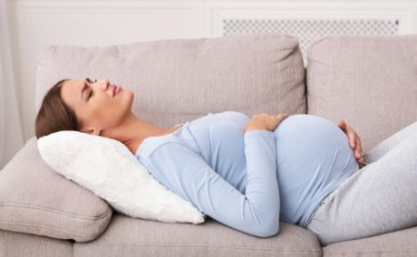 Femeie însărcinată, îmbrăcată cu o bluză bleu și colanți gri, care stă întinsă pe o canapea bej, cu o pernă albă sub cap, și se ține cu mâinile de burtă, ilustrând durerea de ligament rotund în sarcină