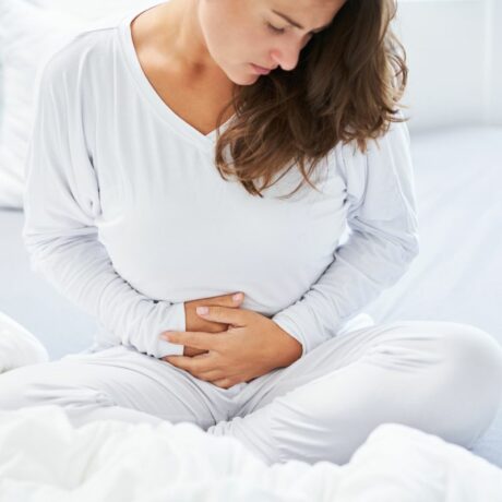 Femeie, îmbrăcată în alb, care își ține mâinile sub abdomen, care stă în pat, cu picioarele încrucișate, ilustrând îngrijirea perineului după naștere