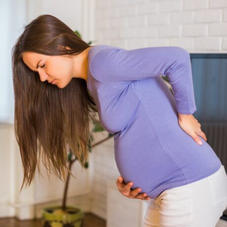 Femeie însărcinată, îmbrăcată cu o bluză mov și pantaloni albi, care stă în living, având în spate un televizor, puțin aplecată, ținând o mână pe spate și una sub burtă, ilustrând înțepăturile vaginale în sarcină
