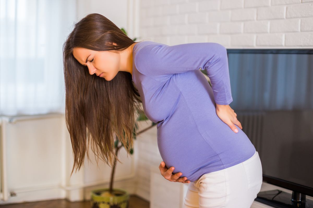 Femeie însărcinată, îmbrăcată cu o bluză mov și pantaloni albi, care stă în living, având în spate un televizor, puțin aplecată, ținând o mână pe spate și una sub burtă, ilustrând înțepăturile vaginale în sarcină