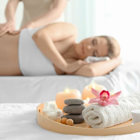 Femeie însărcinată, care stă pe-o parte pe un pat de masaj șoi terapeutul îi masează spatele, iar pe un alt pat de masaj apare o tavă din lemn pe care sunt așezate lumânări, pietre de masaj, niște prosoape mici albe, rulate, și o floare de decor, ilustrând masajul în sarcină
