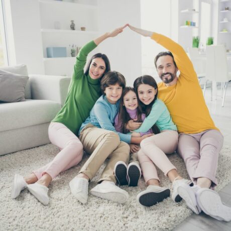Familie fericită cu trei copii, așezată pe un covor bej în living și în spate având o bucătărie open space, ilustrând stilurile parentale