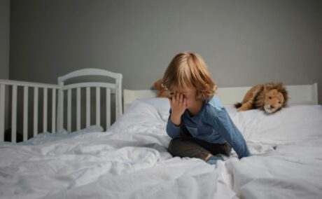 Toddler, care stă în pat de adulți, în spate are un lu din pluș, iar lângă el este lipit un pătuț alb de bebeluși, stă pe o pilotă albă, în timp ce se lovește cu palma peste față, ilustrând toddlerul se lovește