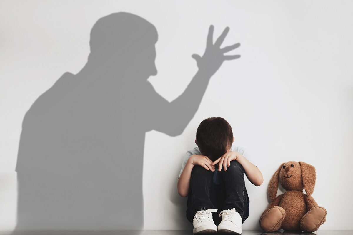 Un copil care stă cu mâinile la față lângă un perete și are umbra unui adult furios deasupra lui
