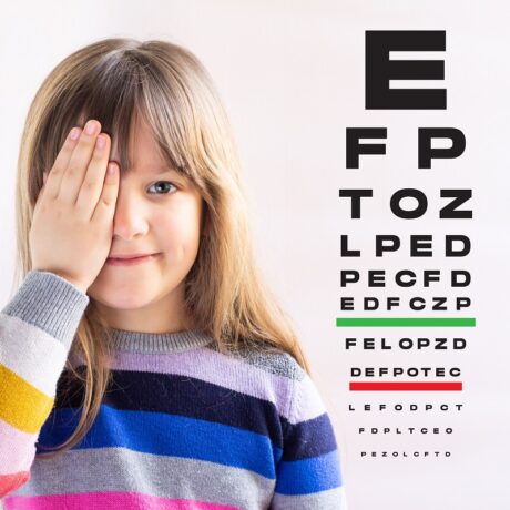 O fetiță care își ține un ochi acoperit și care stă lângă un panou oftalmologic