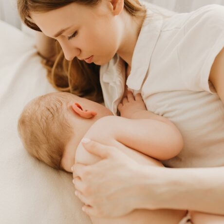 Mamă tânără își alăptează bebelușul întinsă pe pat