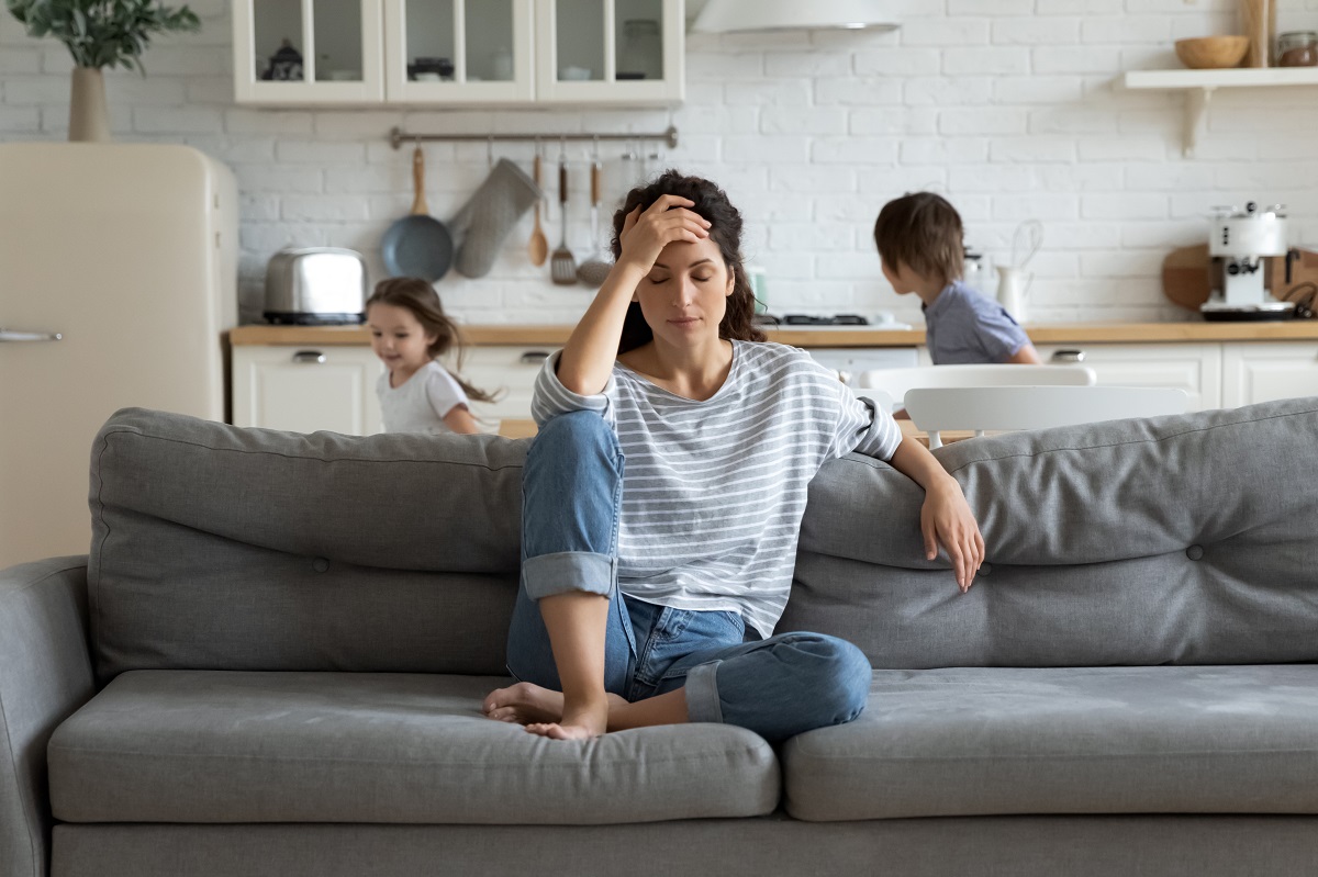 Mama anxioasă care stă pe canapea cu mâna la cap în timp ce copiii aleargă prin casă