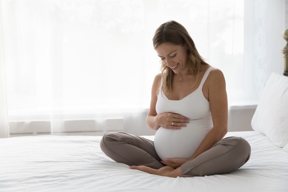Femeie însărcinată stă pe pat cu mâna pe burtă și zâmbește