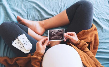 Femeie însărcinată stă în pat și se uită la ecografie cu o pereche de șosete pe picior
