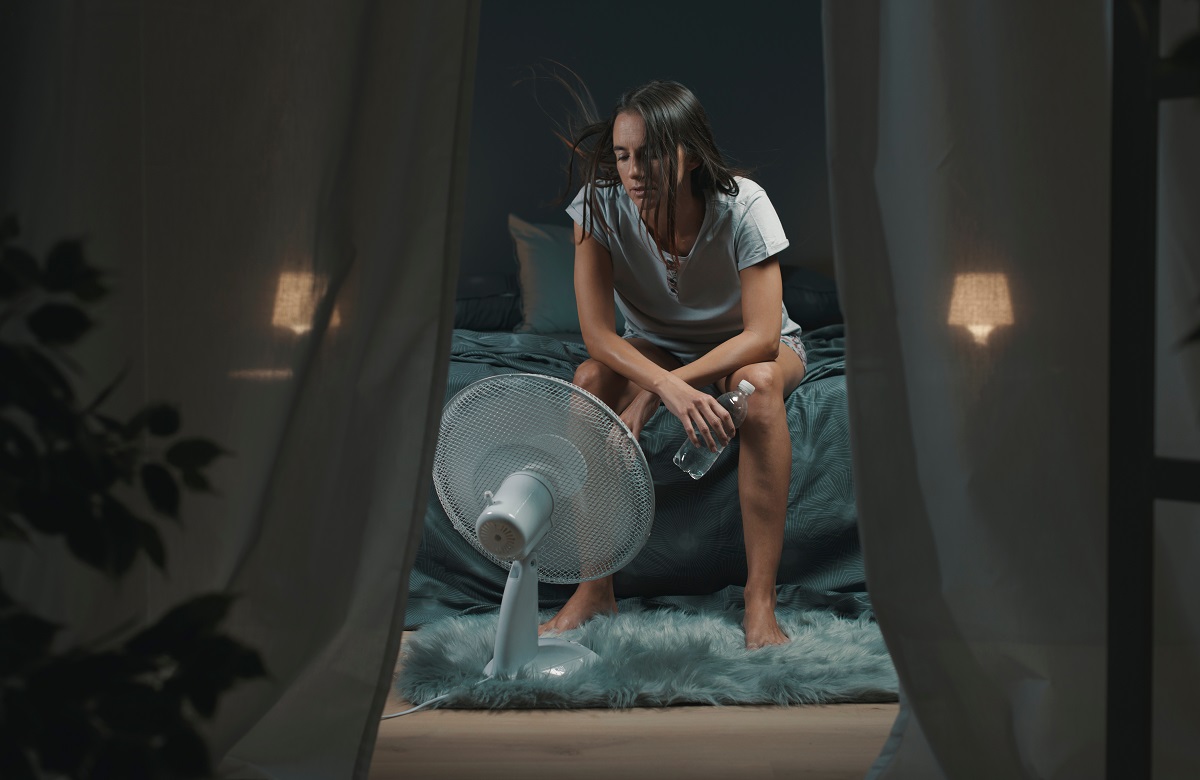 Femeia stă pe marginea patului cu sticla de apă în mână și cu ventilatorul pornit