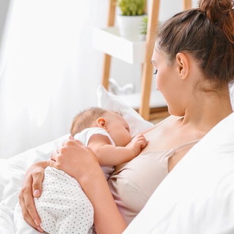 Femeie tânără stă în pat și își alăptează bebelușul